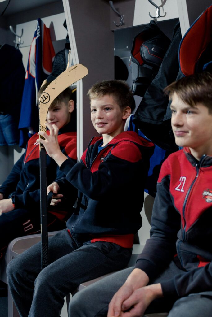 Group of young boys in a locker room preparing for a hockey game, holding gear and sticks.